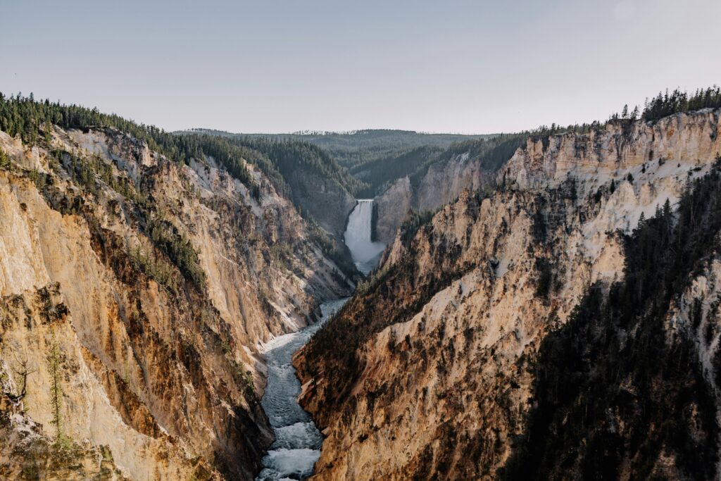 Grand Teton and Yellowstone scenic landscape featuring a waterfall cascading into a river surrounded by rugged cliffs and lush forests, highlighting the natural beauty of Teton Expeditions tours.