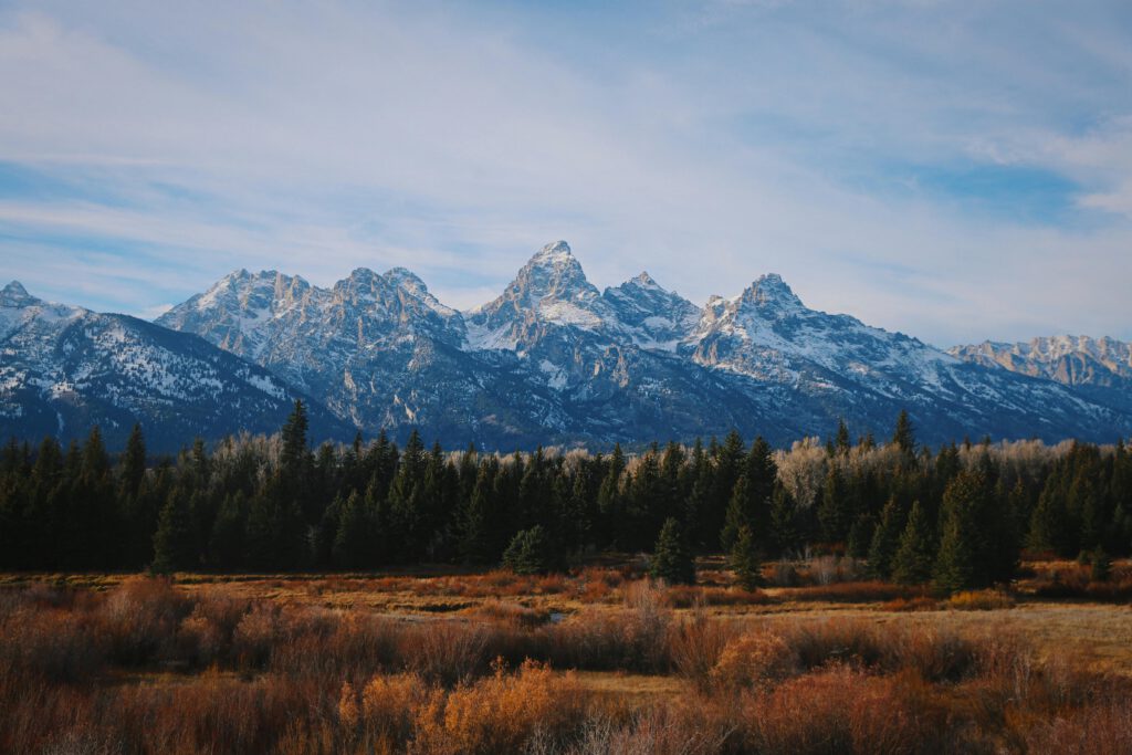 Scenic view of the Teton mountain range with snow-capped peaks, surrounded by evergreen trees and autumn foliage, highlighting the natural beauty of outdoor adventures offered by Teton Expeditions.