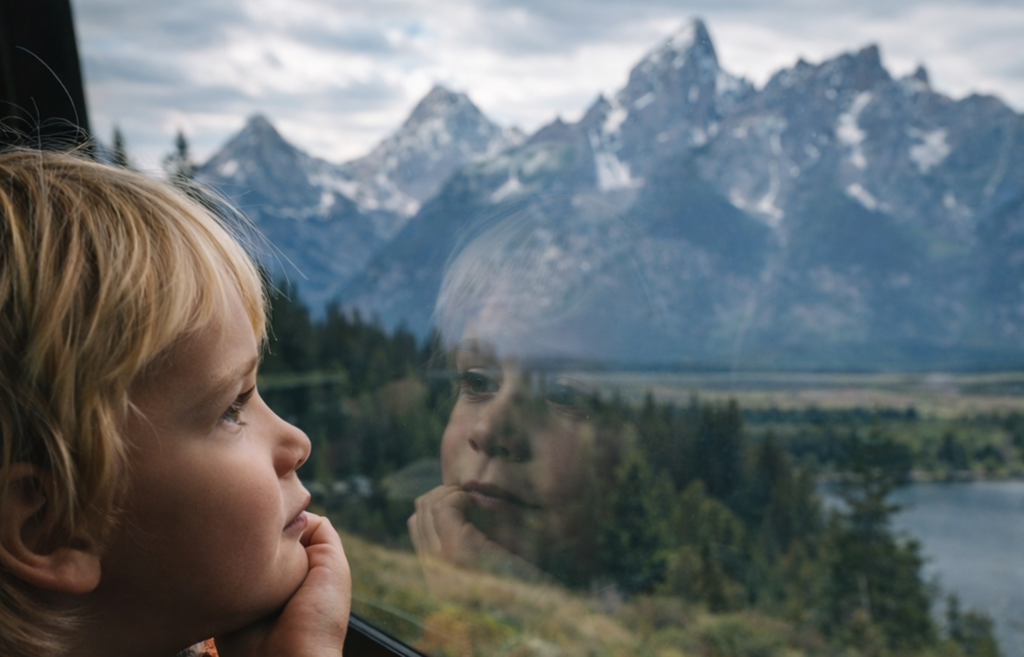 Child gazing out a window at the majestic Teton mountain range, reflecting a sense of wonder and adventure, capturing the essence of scenic tours offered by Teton Expeditions.