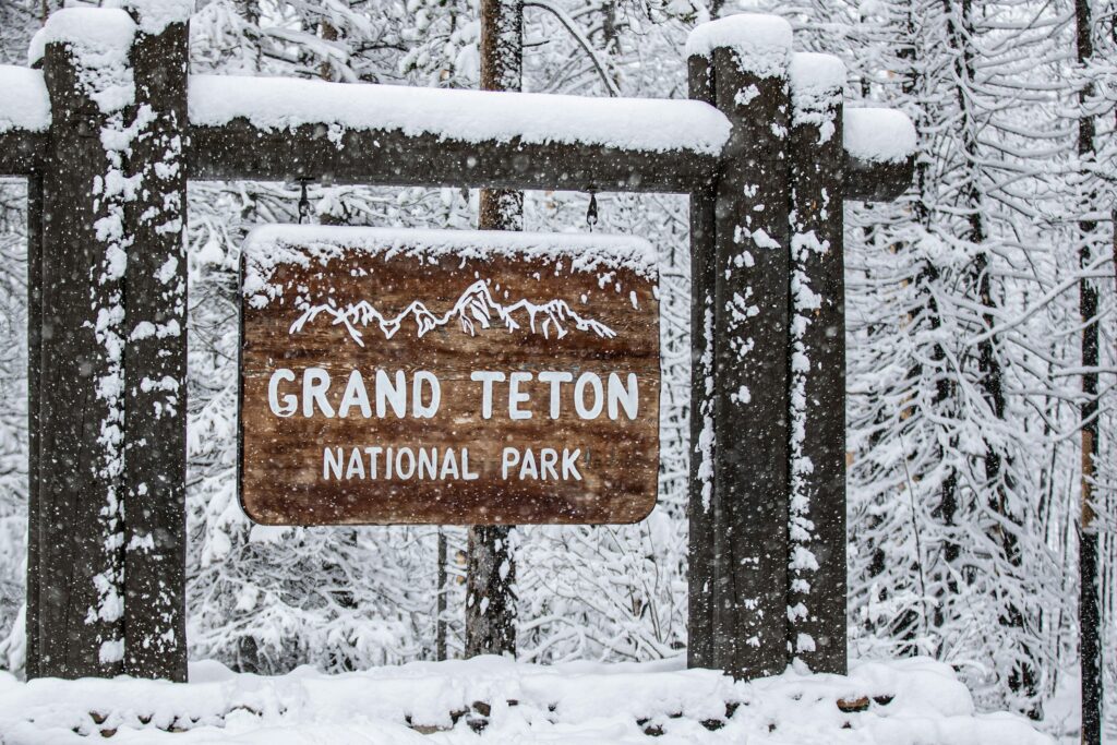 Grand Teton National Park sign covered in snow, surrounded by snowy trees, showcasing winter scenery relevant to Teton Expeditions' scenic tours.