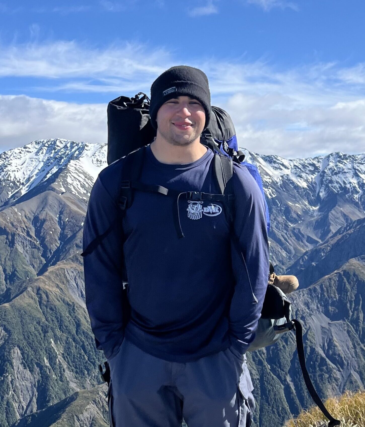 Person wearing a beanie and backpack, standing in front of snow-capped mountains, representing outdoor adventure and guiding experiences in nature.