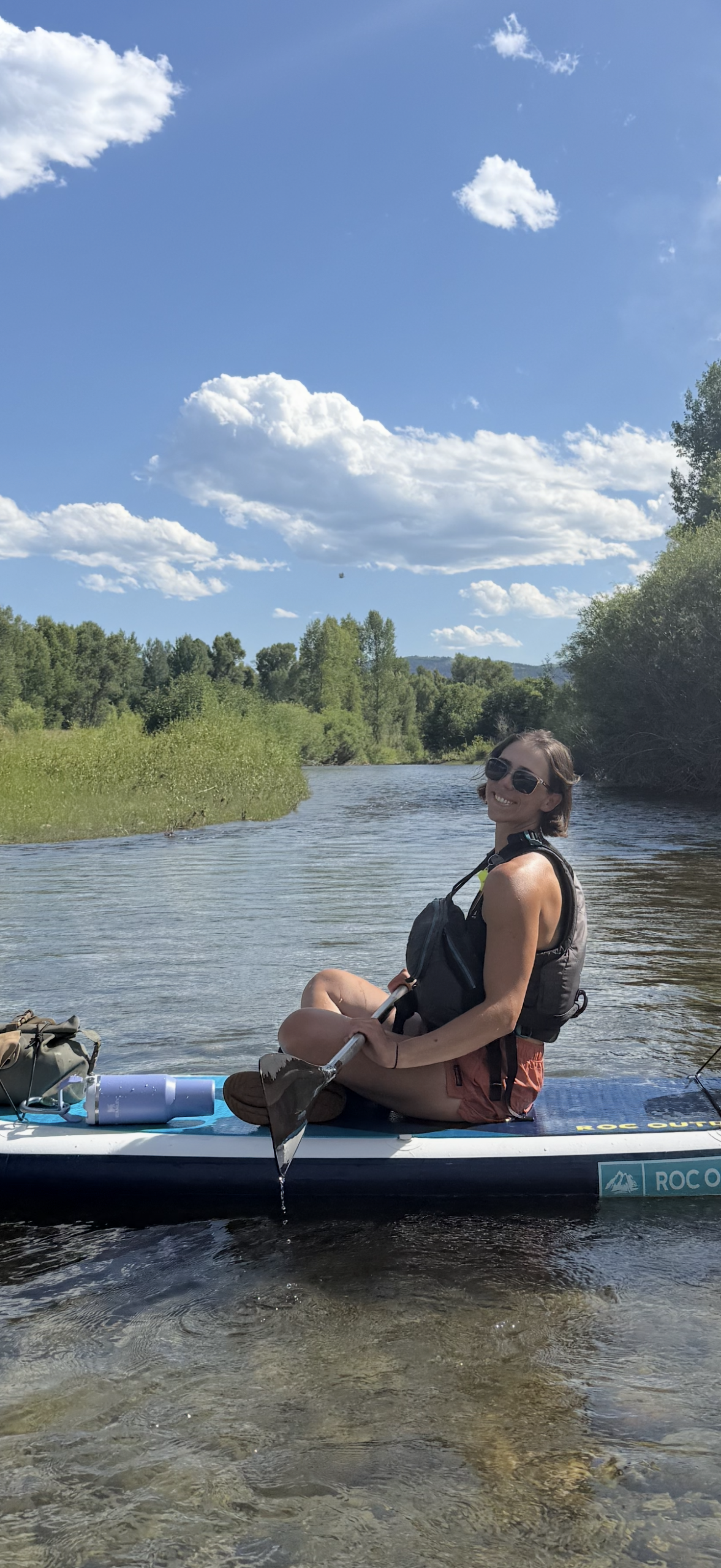 Young woman on a paddleboard in a river, wearing sunglasses and a life jacket, smiling and holding a paddle, surrounded by lush greenery and blue sky with clouds, reflecting outdoor adventure and guiding experiences in the Greater Yellowstone ecosystem.