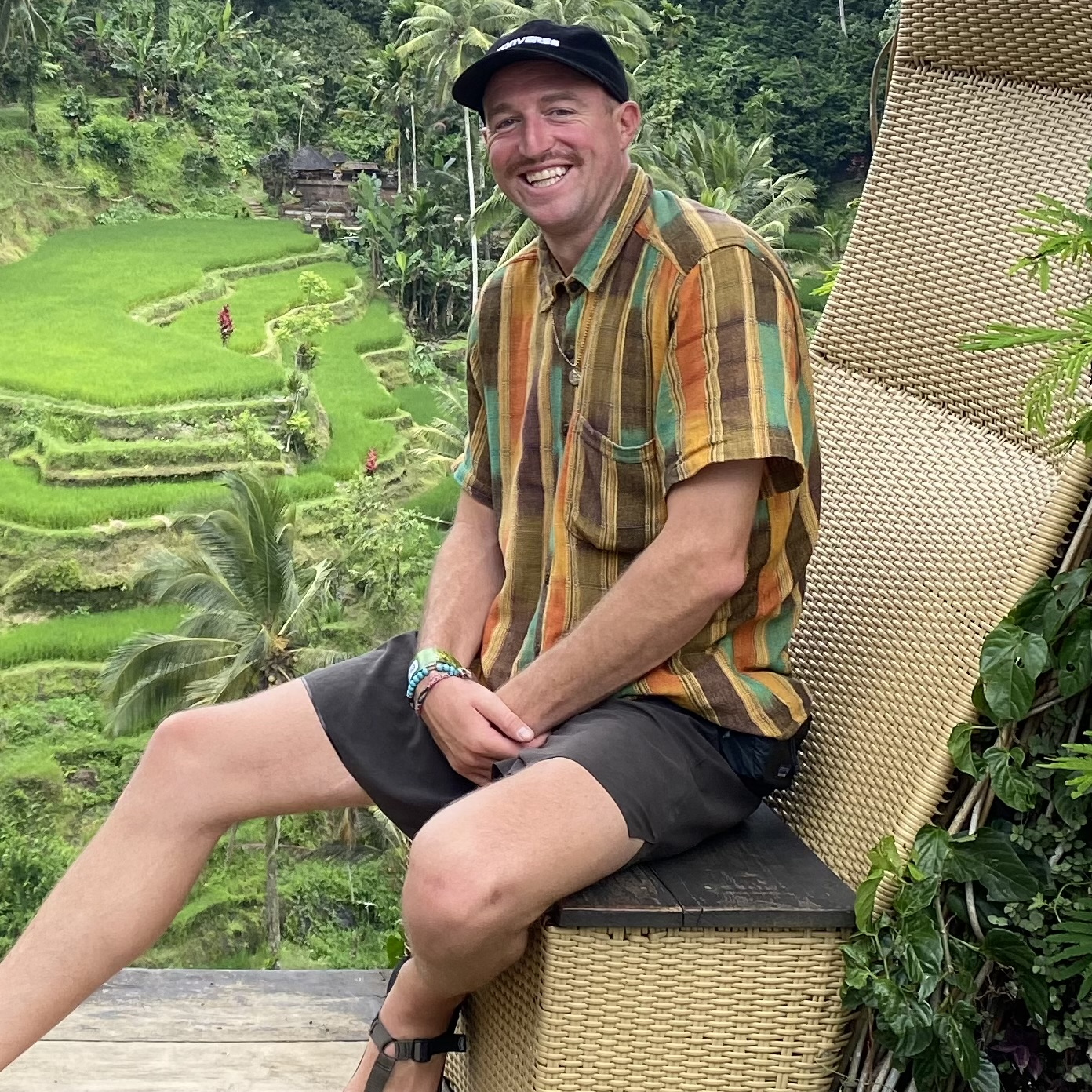 Man sitting on a chair with a colorful striped shirt, smiling against a backdrop of lush green rice terraces and tropical vegetation.